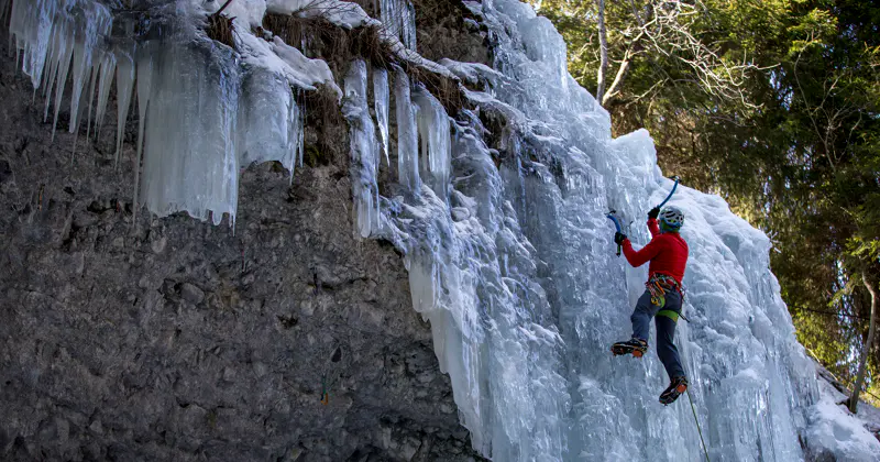 Een persoon in klimuitrusting beklimt een bevroren waterval met behulp van ijsbijlen, omringd door ijspegels en sneeuw, met bomen op de achtergrond.